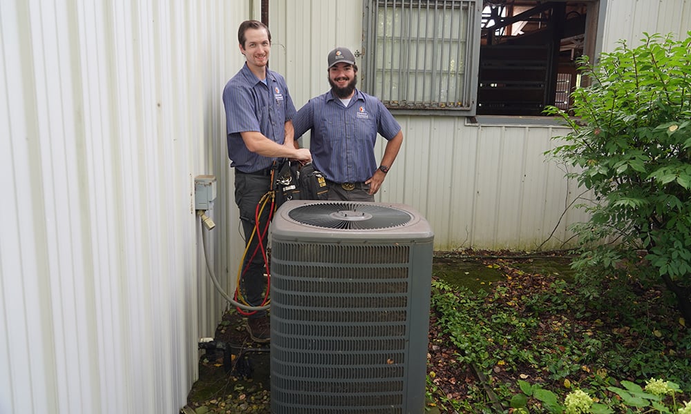 Two HVAC technicians servicing an outdoor AC unit next to a metal building