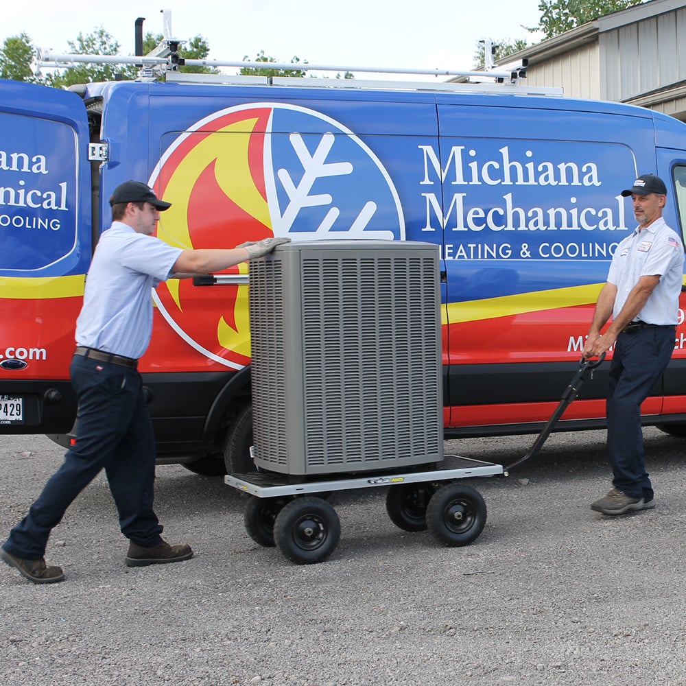Two technicians moving an outdoor AC unit on a wheeled cart in front of branded service van